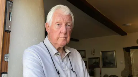 Resident Barrie Atkinson looking at the camera and stood in a doorway. He has his arms by his side.
