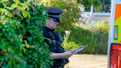 A police officer looks down at a sheert of paper while wearing uniform next to a bush and police van outside Widdecombe's home on Friday.
