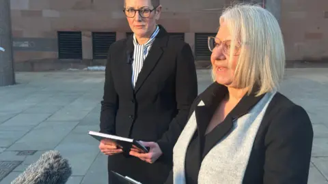 Two women stand outside a court reading statements from clipboards. One has neck length blonde hair and glasses, the other short cropped brown hair and glasses.