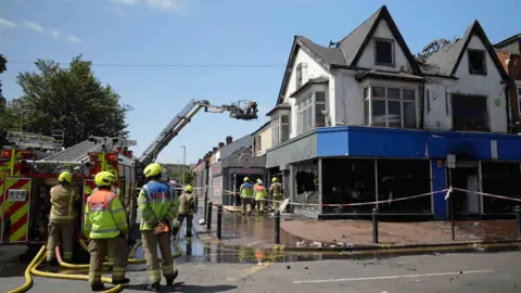 BBC Three fire staff are standing behind a red and yellow fire engine, which is parked in front of a shop. that has blue and black frontage. There is a red and white cordon, and its windows are smashed. There are parts of the roof missing and the first floor is blackened with fire. The fire engine has deployed an aerial ladder which is hovering above the building