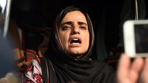 Mahrang Baloch, a woman, is pictured mid-speech with a serious expression. A hand holding a mobile phone aloft, as if to record her speech, is pictured to the right of the frame.