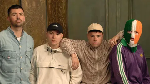 Getty Images Four young men are standing in a room looking at the camera. The man on the left is holding his hands behind his back and wearing a denim top with a white top under it. The two men next to him are wearing caps on their heads and the fourth man is wearing a green, white and orange balaclava over his face.