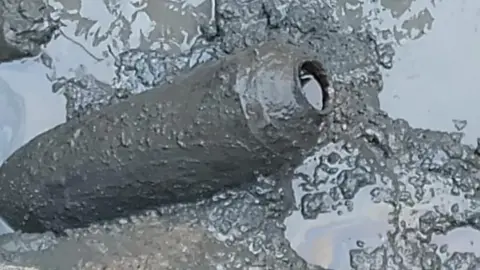 A WWII ordnance device covered in grey mud. The device is cylindrical and has what looks like a hole at one end.
