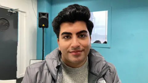 A young man with black hair and a side parting wearing a grey puffer jacket and jumper.