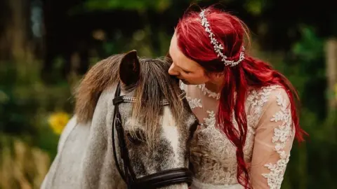 Woman with long red hair in a lace white wedding dress, kissing the head of a brown and grey horse