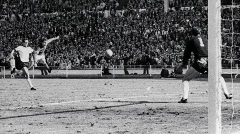 Black and white image shows England's Geoff Hurst in mid-air with his left arm and left leg outstretched as he cracks a shot past German goalkeeper Hans Tilkowski to score the final goal of the World Cup Final against West Germany at Wembley in 1966.