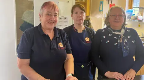 Anne Kemp, Sharman Hoey and Brenda Cunningham smile at the camera behind the counter at Bede's Bakehouse. Anne has short white hair with pink fringe and is wearing a navy blue t-shirt with the black and yellow logo of St Peter's Church. Sherman's brown hair is tied in a pony tail and she is wearing a navy blue apron with the church logo. Brenda has a straight shoulder-length white hair and is wearing a navy blue jumper with embroidered flowers and a scarf. She has red thin-rimmed glasses. 