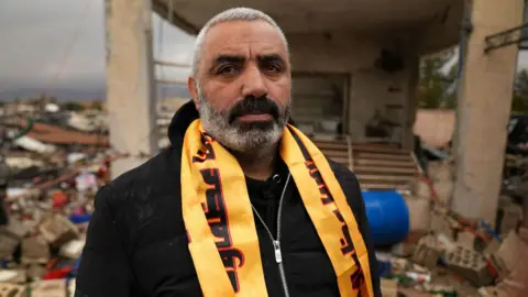 Hassan al-Tahan stands in front of the demolished building with a yellow Hezbollah scarf around his neck