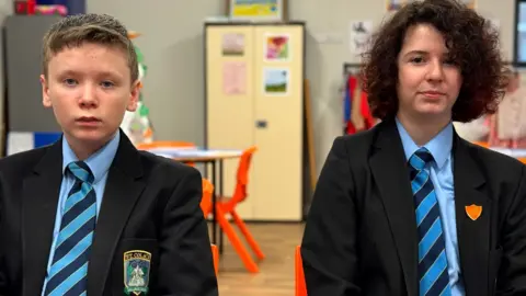 A male and female student are pictured in a classroom. The boy has short blonde hair and the girl has chin-length dark curly hair. They are both dressed in a school uniform - a dark coloured blazer, a light blue shirt, and a blue and yellow striped tie.