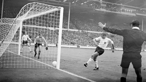 PA Media A black and white photo taken during the World Cup final in 1966. The referee is standing on the right, while a player is kicking the ball into a white net on the left. 