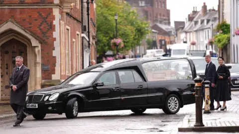 PA Black funeral hearse turns into Crown Street in Bury St Edmunds for Lord Tebbit's funeral. A man in a black coat walks in front with his head bowed. Two mourners follow behind
