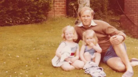 Passion Pictures An old family photo showing Smyth wearing blue shorts and a brown jumper, sitting on the grass beside his two young daughters. It appears to be a warm day, and they are smiling at the camera.