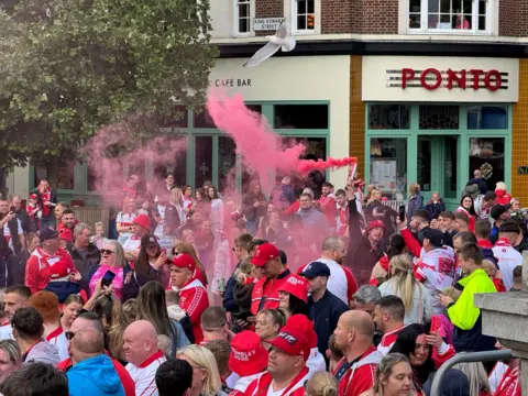 A man holds a flare above his head as it emits red smoke. He is surrounded by a large crowd of people who are wearing red and white clothing.