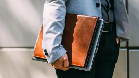 Anonymous man in grey jacket and black formal trousers holds a tan leather document holder under his harm.