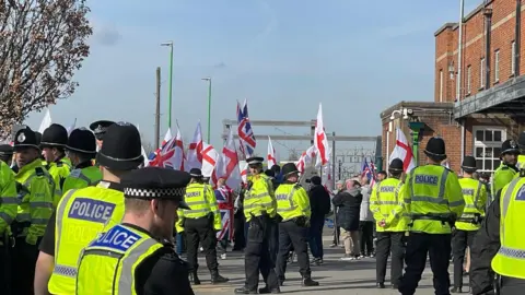 BBC Many police officers with their backs to camera facing a group of protestors holding England flags and Union Jacks