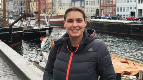 Family Handout A woman with brown hair tied-up is wearing a black Adidas raincoat with an orange zip and smiling. She appears to be standing on a harbourside with boats behind her and a row of colourful houses.