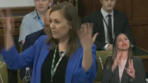 MP Jen Craft, wearing a blue jacket and black top with shoulder-length hair, uses sign language as part of a House of Commons debate