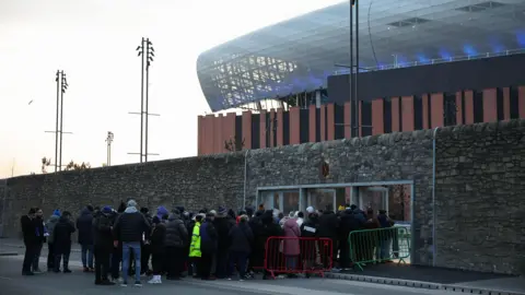 EPA Fans queue to enter Everton's Bramley Moore Dock stadium in Liverpool wearing coats and hats. The blue and brown stadium stands in the background, behind a grey stone wall, which houses a number of turnstiles