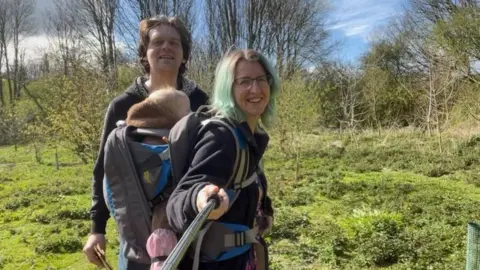 Daisy MacDermott A woman with short, dyed green hair carries on a baby in a rucksack on her back. A man with brown hair stands beside her. In the background, a grassy field and trees can be seen.