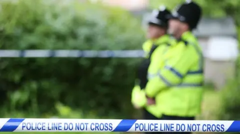 Two police officers in bright yellow jackets and helmets stand behind police tape. 