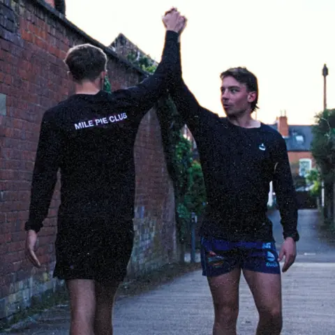 Pritchard Brothers The same two men on a city street. One is walking towards the camera and the other is walking away as they raise their hands for a high-five. They are both wearing shorts and long-sleeved black t-shirts. One of the back of the one walking away is white lettering reading "MILE PIE CLUB".