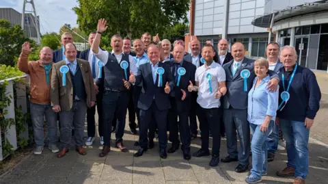 A group of Reform politicians wearing light blue rosettes and smiling.