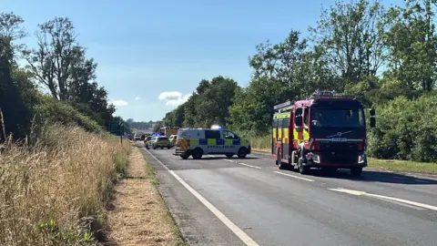 A road with trees on either side and several police and fire vehicles parked on it.