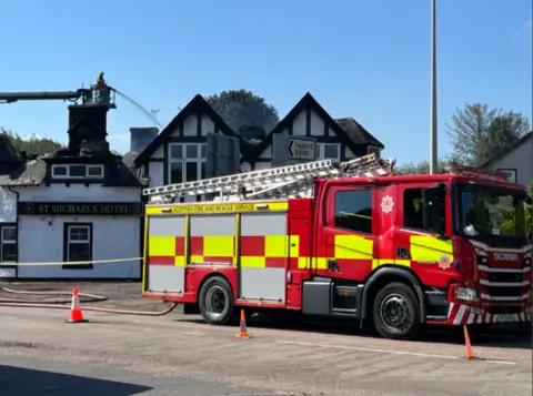 A Scottish Fire and Rescue Service fire engine is parked outside St Michaels Hotel, where firefighters on an aerial platform tackle damage to the roof, with hoses, cones and a safety cordon visible.