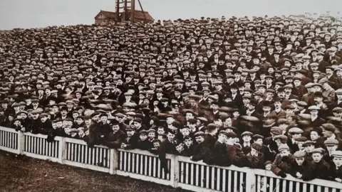 Hull City Supporters' Trust A black and white photo of the Spion Kop. Hundreds of football fans, many wearing flat caps, stand behind a wooden fence