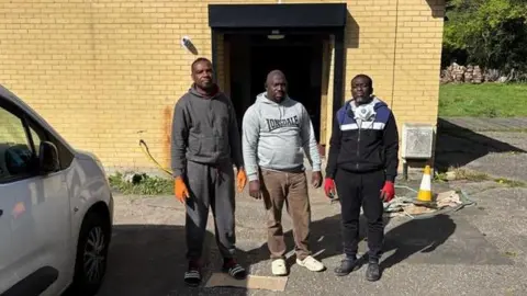 Daniel Berkoh-Gyamfi Three men standing in front of the church building, they are all wearing gloves and hoodies. There is a white car on the side and orange and white cones. The men look upset. 