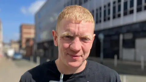 Ewan Welsh, a man wearing a black t-shirt, is looking at the camera. He is stood on a street in Stoke-on-Trent city centre.