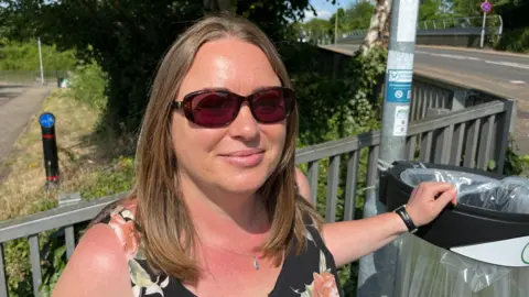 John Devine/BBC Samantha Dalton, 48, has brown/light blonde shoulder length hair parted in the middle of her head. She has dark glasses and a silver chain around her neck. She is wearing a dark floral sleeveless dress and has her hand on the rim of a public bin. Behind her are railings and roadways.