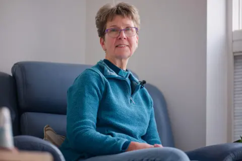 Laurence Cawley/BBC Judith Gibbons sits in a blue chair in her living room. She is wearing a blue jumper, a blue sparkly dog collar and has purple-rimmed glasses