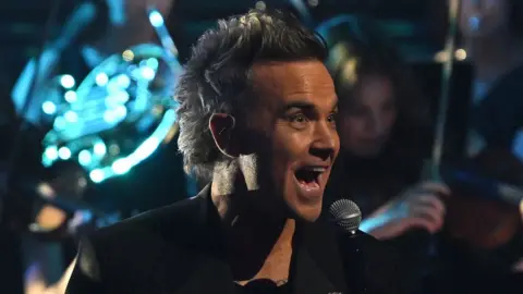 Robbie Williams smiles while singing on stage with a backing band in dark clothing.