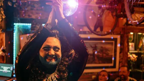 Sarah Hewitt A drag queen with a blue beard claps above their head