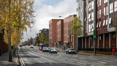 Belfast City Council An artist's impression of the new apartments at the site of Havelock House. There are trees on the left of the road and a brown apartment building and a grey office block on the right. Cars and a bus travel down the road. 