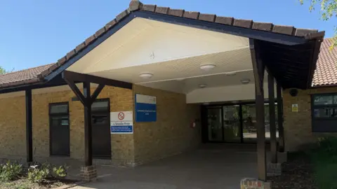 Elliot Deady/BBC Outside a one-storey hospital building. It has an arched porched area welcoming patients to its glass doors. There is blue sky above.