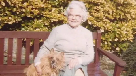Mabel sitting on a wooden bench with her small terrier-type dog with long light brown fur. Mabel has curly white short hair and glasses, and is wearing a cream jumper and light green long skirt. There is a hedge behind her. 