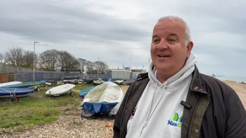 Ian Pickard, who co-founded Mankind, smiling. He is standing on a beach in Eastbourne with small boats docked behind him.