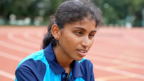 BBC Head shot of Rakshitha with long dark hair tied in a pony tail, red and gold stud earrings and a shirt made from light blue and dark blue fabric. There is a reddish-brown running track in the background.   