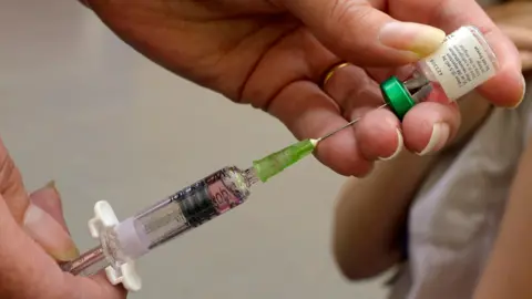 A pair of hands holding a syringe which has it's needle in a vaccine vile.