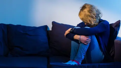 A woman with curly blonde hair sat on a navy blue sofa. She has her head down and arms around her legs.