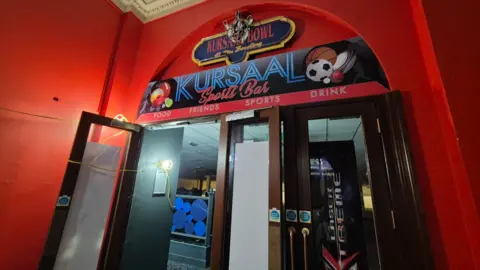 Stuart Woodward/BBC The entrance to the bowling alley, surrounded by a red-painted wall. KURSAAL SPORTS BAR is printed above the doors.