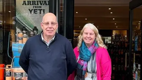 Family photo Andrew Fuller, wearing a dark blue jumper with zipped top and shirt underneath, next to his sister Liz, wearing a bright pink jacket and pink and blue patterned scarf. Both are smiling as they stand outside the front of their shop, SC Fuller, with outdoor merchandise visible