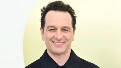 Rhys on a red carpet, wearing a black shirt. He is clean-shaven with short dark hair, and is standing against a pale yellow backdrop. 