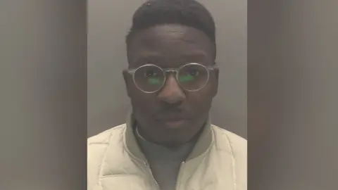 The mugshot of a man wearing transparent glasses and white jacket over a grey jumper. 