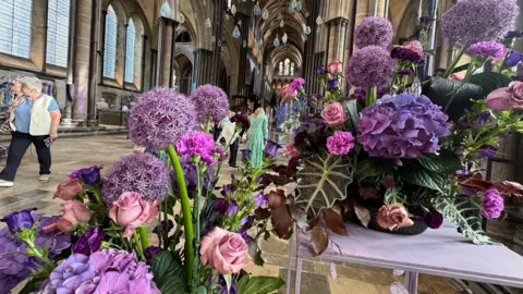 An arrangement of purple and pink flowers on two white tables. You can see people in the hall walking around looking at things in the background.