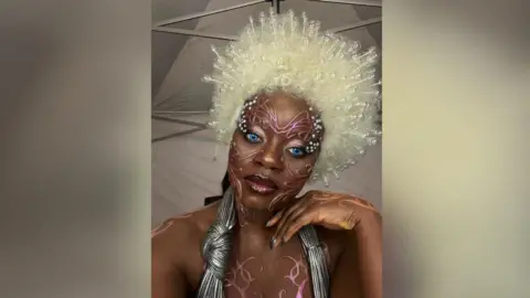 Winnie Ama A person in bold avant‑garde makeup with bright blue eyes, a large white beaded wig, and metallic costume accessories, standing in a tent-like backstage setting