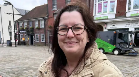 Natalie is wearing brown and black patterned classes and a beige coat with a faux-fur lined hood.