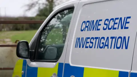 A close-up of a West Yorkshire Police van. It is mainly white, with the words "Crime Scene Investigation" written on the side in blue. There is a gate and a field in the background.